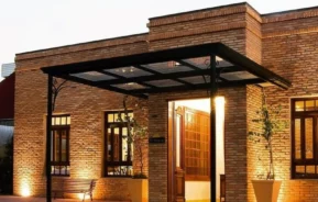 Prédio de tijolos de um andar com janelas grandes, iluminação quente e um dossel de vidro e metal na entrada. Dois grandes vasos de plantas e um banco estão colocados perto da entrada. Céu noturno ao fundo.