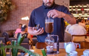 Imagem de um homem de camisa preta preparando café usando um sifão em uma cafeteria, com vapor saindo do dispositivo, para ilustrar matéria sobre as cafeterias em Florianópolis. As pessoas sentam e conversam em um fundo aconchegante e suavemente iluminado. Plantas verdes são visíveis em primeiro plano.