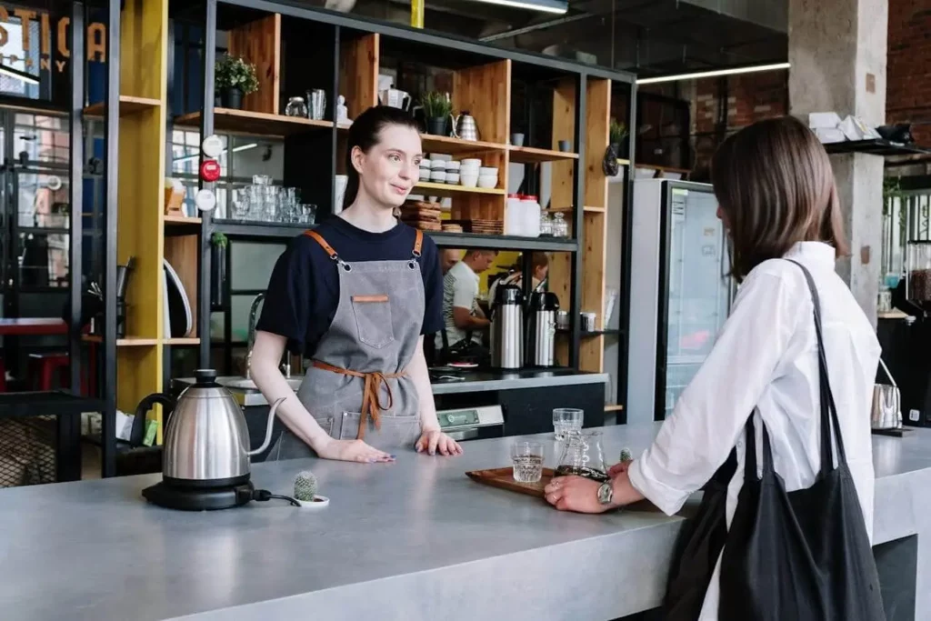 Imagem de uma barista com avental cinza está atrás do balcão de uma cafeteria, sorrindo e conversando com um cliente que está vestindo uma camisa branca e carregando uma bolsa preta, para ilustrar matéria sobre as cafeterias em Florianópolis bem avaliadas. O balcão tem uma chaleira, copos e um pequeno vaso de plantas.