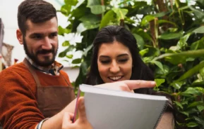 Imagem de duas pessoas em pé entre plantas verdes, sorrindo e olhando juntas para um caderno, com uma pessoa apontando para algo na página, para ilustrar matéria sobre o CNIR.