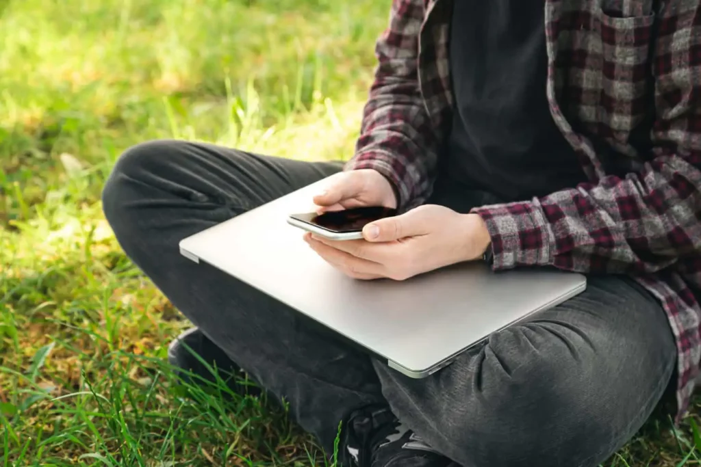 Imagem de uma pessoa sentada de pernas cruzadas na grama, segurando um smartphone com as duas mãos e com um laptop fechado no colo. Somente o tronco, as mãos e as pernas estão visíveis.