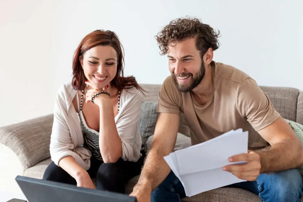 Imagem de um casal sorridente sentado em um sofá olhando para um notebook. O homem segura várias folhas de papel e a mulher apoia o queixo em sua mão. Eles parecem comprometidos e felizes