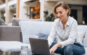 Imagem de uma mulher vestindo camisa floral e calça jeans sentada em um sofá, sorrindo enquanto usa um notebook, para ilustrar matéria sobre consórcio imobiliário para quem já tem entrada. Um copo de água está sobre a mesa à sua frente em uma sala de estar moderna e bem iluminada.