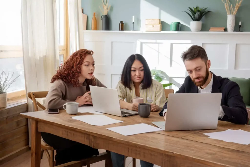 Imagem de três pessoas sentadas em uma mesa de madeira com notebooks, papéis e canecas, trabalhando e discutindo juntas em uma sala moderna e iluminada com plantas e prateleiras ao fundo.