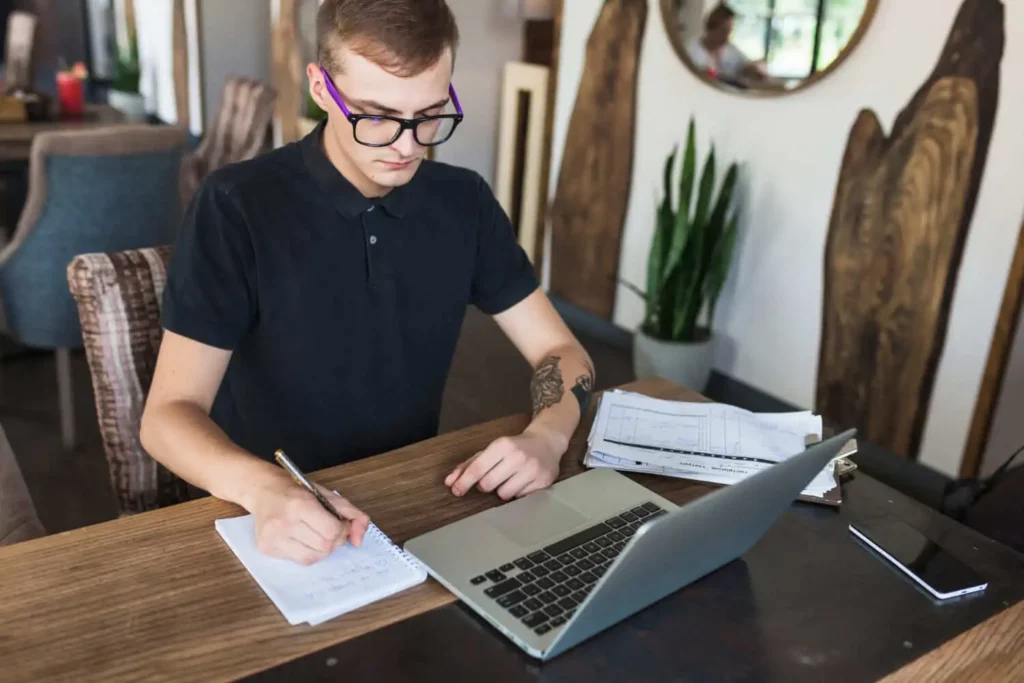 Imagem de uma pessoa usando óculos e uma camisa polo preta escreve em um caderno enquanto trabalha em um laptop em uma mesa de madeira. Papéis e um smartphone estão próximos, e plantas e decoração em madeira são visíveis ao fundo.