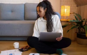 Imagem de uma mulher sentada de pernas cruzadas no chão com um notebook no colo, usando uma calculadora, para ilustrar matéria sobre o reajuste no consórcio. Papéis, uma caneta e uma xícara estão no chão ao seu redor. Um sofá, uma planta e uma lâmpada acesa estão ao fundo.