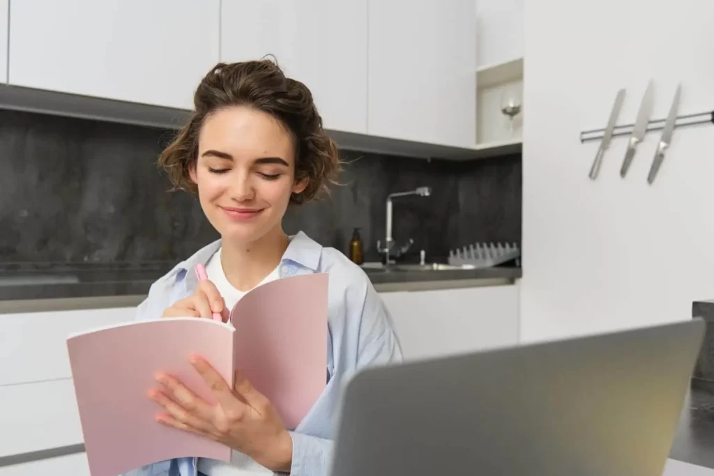 Imagem de uma mulher sentada em uma cozinha moderna, sorrindo enquanto escreve em um caderno rosa. Ela está olhando para o caderno, com um laptop aberto à sua frente no balcão.