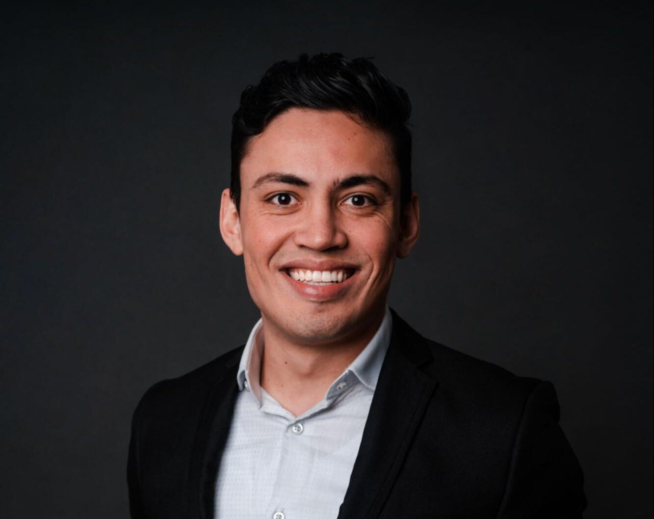 Um homem de cabelos escuros, vestindo uma camisa branca de colarinho e um blazer preto, sorri para a câmera contra um fundo escuro simples.