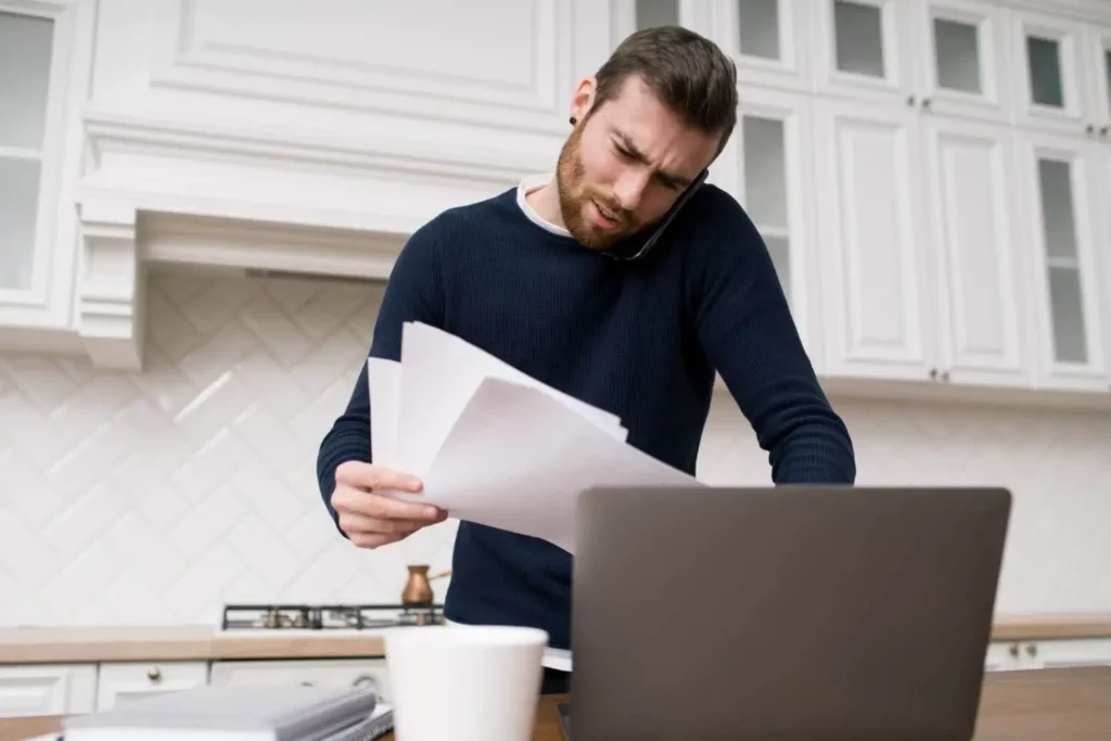 Imagem de um homem em uma cozinha, segurando papéis e falando ao telefone enquanto olha para um laptop. Ele parece concentrado e preocupado. Uma xícara de café e mais papéis estão sobre o balcão à sua frente