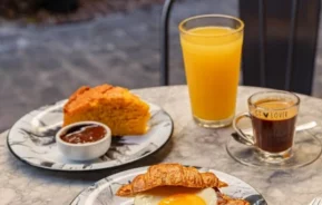Um café da manhã servido em uma mesa de mármore com um sanduíche de croissant coberto com um ovo frito, uma fatia de bolo com geleia, um copo de suco de laranja e uma xícara de café expresso, ao ar livre.