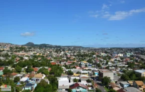 Uma vista panorâmica de uma paisagem urbana com várias casas, árvores verdes e colinas distantes sob um céu azul claro com nuvens dispersas.