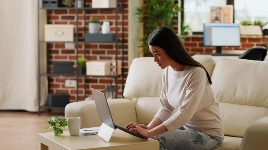 Imagem de uma mulher sentada em um sofá bege, trabalhando em um laptop em uma mesa de centro com uma caneca e uma pequena planta. A sala tem paredes de tijolos, prateleiras e plantas de interior, criando uma atmosfera aconchegante de home office.