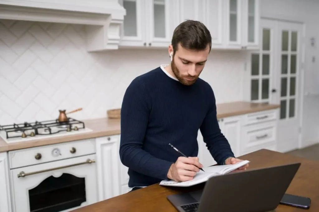 Imagem de um homem de barba está em pé em uma cozinha moderna, escrevendo em um caderno com um lápis enquanto olha para um laptop no balcão de madeira. A cozinha tem armários e eletrodomésticos brancos.