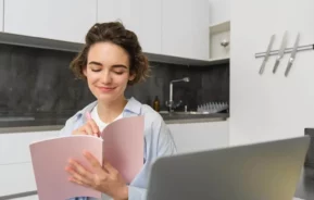 Imagem de uma jovem mulher sentada em uma cozinha moderna, sorrindo enquanto escreve em um caderno rosa. Um laptop aberto está à sua frente, no balcão, e várias facas estão montadas na parede ao fundo.