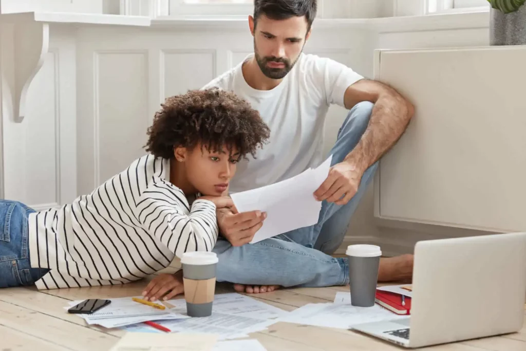 Imagem de um casal sentado no chão, concentrado em papéis e documentos espalhados ao seu redor. Eles olham os papéis juntos, com um laptop e xícaras de café por perto.