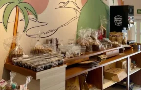 Foto que ilustra matéria sobre cafeterias em Santos mostra uma exposição de produtos de padaria e chocolates embalados variados em prateleiras de madeira em uma padaria, com um mural colorido de palmeiras e uma montanha na parede atrás no Bossa Café & Bistrô.