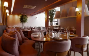 O interior do restaurante é elegante, com cadeiras e sofás lilás, mesas com copos e talheres, iluminação suave nas paredes e vasos de plantas, criando uma atmosfera moderna e aconchegante.