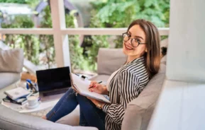 Imagem de uma mulher usando óculos e camisa listrada sentada em um sofá, sorrindo para a câmera enquanto escreve em um caderno. Um laptop, uma xícara e papéis estão sobre a mesa ao lado dela. Grandes janelas mostram a vegetação do lado de fora.
