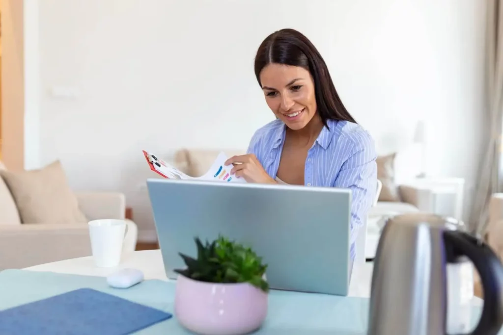Imagem de uma mulher sentada em uma mesa trabalhando em um laptop, sorrindo enquanto olha para um documento com gráficos coloridos. Uma caneca de café, um vaso de planta e uma chaleira estão sobre a mesa em uma sala iluminada e aconchegante.