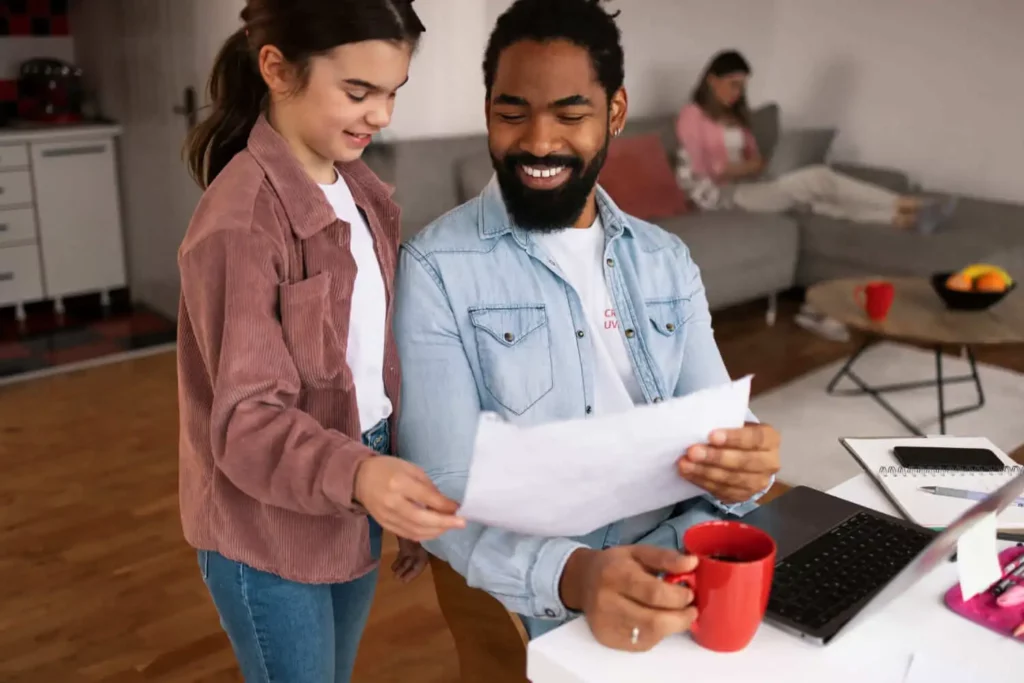 Imagem de um homem sentado em uma escrivaninha com um laptop e uma caneca vermelha enquanto examina papéis ao lado de uma criança. Ao fundo, uma mulher está sentada em um sofá, usando um laptop. Eles estão em uma sala de estar aconchegante e iluminada.