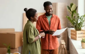 Imagem de duas pessoas, um homem e uma mulher, em pé em um ambiente interno de uma casa, sorrindo e olhando para uma folha de papel. Há caixas de papelão e plantas ao redor delas.