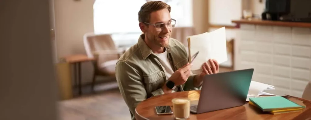 Imagem de um homem sentado em uma mesa redonda com um laptop, um smartphone, livros e um café. Ele sorri enquanto segura um caderno e uma caneta, parecendo participar de uma chamada de vídeo ou de uma reunião on-line em um ambiente interno aconchegante.