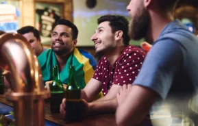 Imagem de quatro homens sentados em um bar, assistindo a um jogo com entusiasmo. Dois deles seguram garrafas de cerveja, um está envolto em uma bandeira verde, amarela e azul, e o grupo parece estar imerso em um ambiente animado.