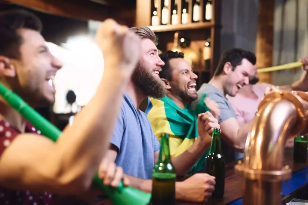 Imagem de um grupo de homens animados assistindo a um jogo em um bar, segurando garrafas de cerveja e vestindo roupas casuais. Um deles tem uma bandeira sobre os ombros, e todos sorriem entusiasmados no balcão.