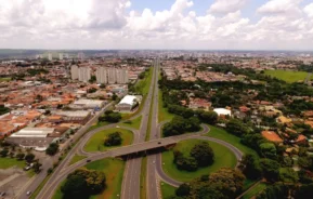 Vista aérea de uma cidade com uma importante rodovia atravessando o centro, viadutos, prédios residenciais ao redor, áreas verdes e árvores espalhadas sob um céu parcialmente nublado.