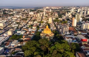Vista aérea de uma cidade com um grande edifício de cúpula amarela cercado por árvores em primeiro plano e muitos prédios residenciais ao fundo, sob um céu limpo.