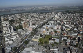 Vista aérea de uma área urbana densamente construída, com inúmeros prédios, ruas e áreas verdes espalhadas sob um céu enevoado, com um rio distante e a paisagem urbana visíveis no horizonte.