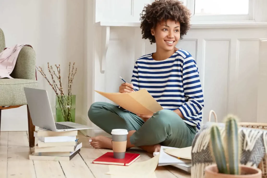 Imagem de uma jovem sentada no chão com papéis, um laptop, livros e café, sorrindo enquanto trabalha. Ela usa uma blusa listrada e está cercada por uma decoração que inclui plantas e uma cesta trançada.