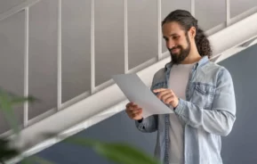 Imagem de um homem de cabelo comprido preso em um rabo de cavalo, vestindo uma camisa jeans clara por cima de uma camiseta branca, sorri enquanto lê uma folha de papel dentro de casa, em pé perto de uma escada, para ilustrar matéria sobre consórcio para comprar imóvel na planta com desconto.