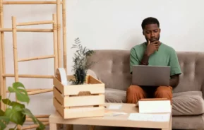 Imagem de um homem sentado no sofá, olhando pensativo para o seu notebook, para ilustrar matéria sobre consórcio ou empréstimo. Uma caixa de madeira com uma planta, alguns papéis e uma caixa de papelão estão sobre a mesinha de centro à sua frente.