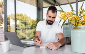 Imagem de um homem escrevendo em um caderno em uma mesa onde há um notebook, uma caneca de café e um vaso com flores amarelas, com vegetação visível através da janela de vidro, para ilustrar matéria sobre a taxa de administração do consórcio.